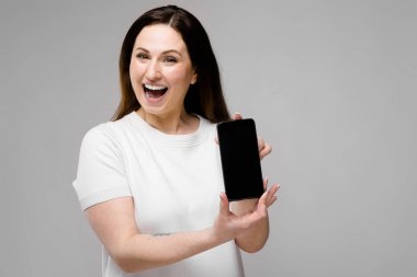 Farklı ifadeler gri duvar ve gösteren boş ekran smartphone üzerinde gösterilen basit beyaz t-shirt uzun lüks saçlı güzel curvy esmer kadın