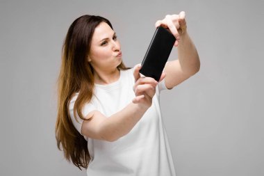 Farklı ifadeler Studio gri duvar selfie onun Smartphone için poz sırasında gösterilen basit beyaz t-shirt uzun lüks saçlı güzel curvy esmer kadın.