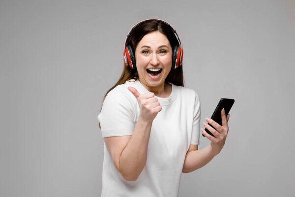 Красивая брюнетка с длинными роскошными волосами в простой белой футболке наслаждается музыкой со смартфоном и наушниками, показывая различные выражения на серой стене в студии
.