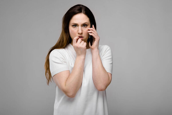 Beautiful curvy brunette woman with long luxurious hair in simple white t-shirt showing different expressions on grey wall while talking on the phone.