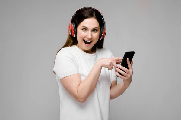 Красивая брюнетка с длинными роскошными волосами в простой белой футболке наслаждается музыкой со смартфоном и наушниками, показывая различные выражения на серой стене в студии
.