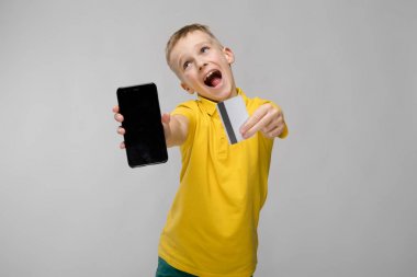 Sevimli sarışın beyaz varoluş çocukta farklı ifadeler gösterilen ve beyaz duvar Studio smartphone ile kredi kartı tutan parlak t-shirt.