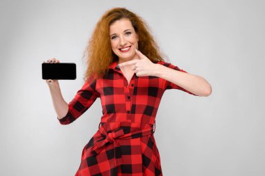 Beyaz duvar Studio farklı duygusal ifadeler gösterilen kırmızı kareli elbiseli güzel kıvırcık saçlı Kafkas kadın. Gülümseyen ve boş smartphone gösterilen.