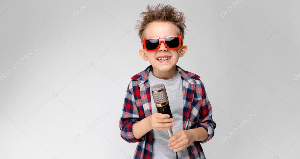 Nice preschooler caucasian boy in casual outfit posing in party sunglasses with microphone and showing different expressions on white wall in studio.