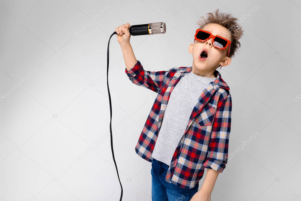 Nice preschooler caucasian boy in casual outfit posing in party sunglasses with microphone and showing different expressions on white wall in studio.