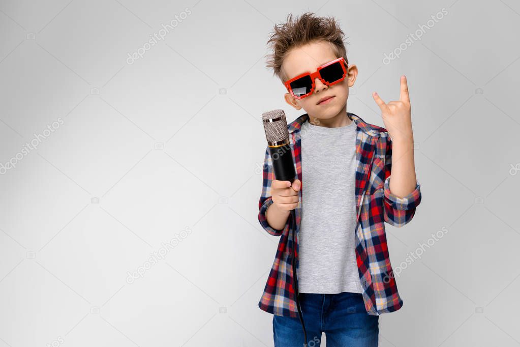 Nice preschooler caucasian boy in casual outfit posing in party sunglasses with microphone and showing different expressions on white wall in studio.