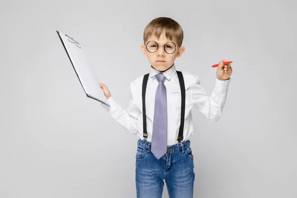 灰色の背景にクリップボードを保持しているメガネで愛らしい深刻な自信を持って小さな男の子の肖像画 — ストック写真