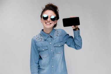 Topuz saç modeli Güneş gözlüğü ile gri izole boş perde smartphone gösterilen genç esmer beyaz kızla
