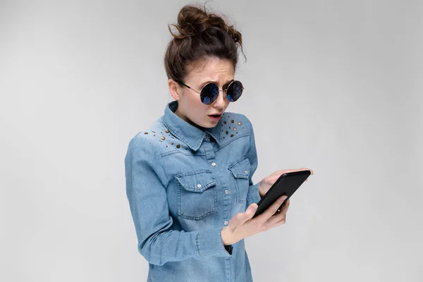 年轻的黑发白种女孩与包子的发型在太阳镜使用智能手机的灰色隔离 — 图库照片
