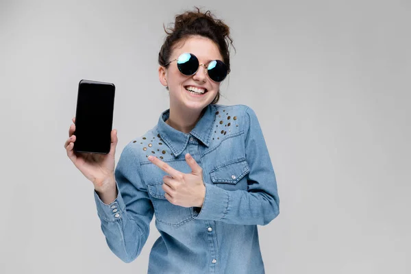 年轻的黑发白种女孩与包子的发型在太阳镜显示智能手机与空白屏幕上的灰色隔离 — 图库照片