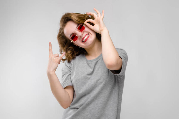 Portrait of beautiful redhead happy young woman in red sunglasses showing rock sign on gray background