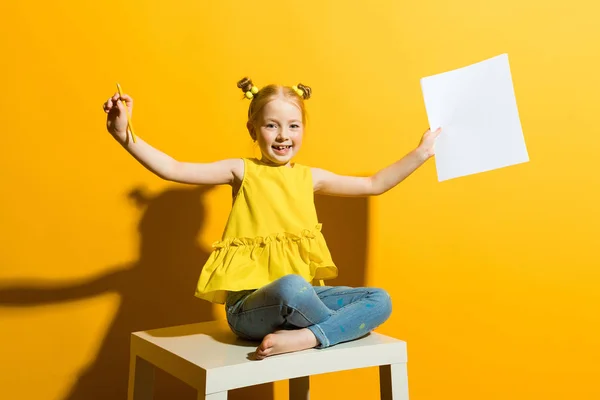 Το κορίτσι με τα κόκκινα μαλλιά σε κίτρινο φόντο. Όμορφη κοπέλα είναι κάθεται στο τραπέζι και να κρατά στα χέρια ένα μολύβι και ένα λευκό φύλλο. — Φωτογραφία Αρχείου