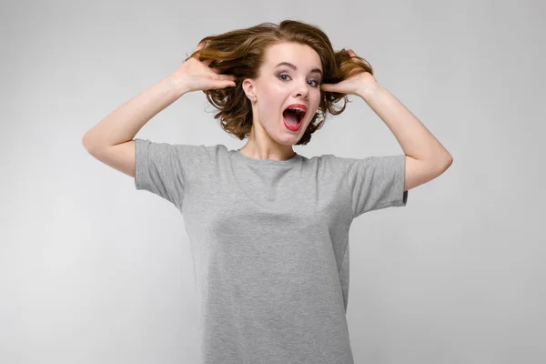 Charming young girl in a gray T-shirt on a gray background. The girl holds her hands behind her head and screams — Stock Photo, Image