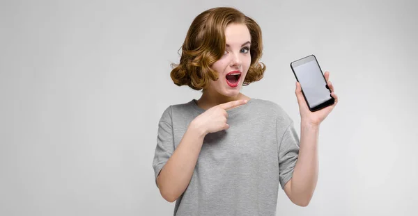 Charming young girl in a gray T-shirt on a gray background. A girl is holding a phone in her hand — Stock Photo, Image