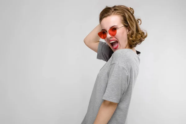 Encantadora joven con una camiseta gris sobre un fondo gris. Chica feliz en gafas rojas. La chica está medio despierta. — Foto de Stock