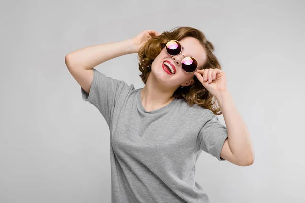 Charmantes junges Mädchen in einem grauen T-Shirt vor grauem Hintergrund. fröhliches Mädchen mit runden Gläsern — Stockfoto