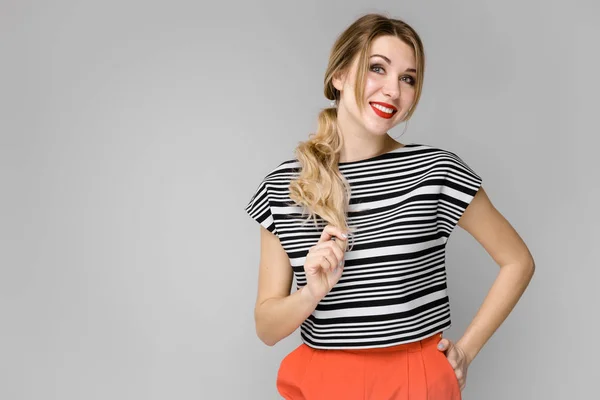 Menina loira jovem atraente em blusa listrada sorrindo segurando seu cabelo em pé no fundo cinza — Fotografia de Stock