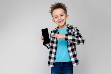 Yakışıklı bir çocuk bir ekose gömlek, mavi tişört ve kot gri bir arka plan üzerinde duruyor. Çocuk bir telefon tutarak