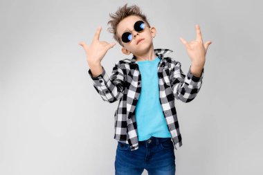 mavi tişört ve kot gri bir arka plan üzerinde duruyor. Oğlan yuvarlak gözlük giyiyor. Kızıl saçlı çocuk bir rocker keçi gösterir