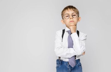 Beyaz bir gömlek, pantolon askısı, kravat ve ışık kot büyüleyici bir çocuk gri bir arka plan üzerinde duruyor. Çocuğun çenesine yumruk üzerinde koymak.
