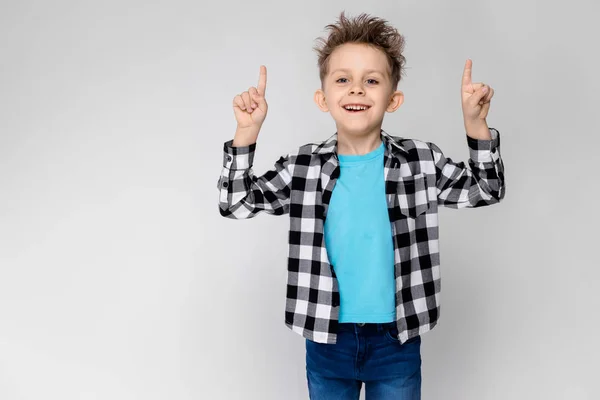 Um menino bonito em uma camisa xadrez, camisa azul e jeans fica em um fundo cinza. Rapaz sorrindo e mostrando os polegares para cima — Fotografia de Stock