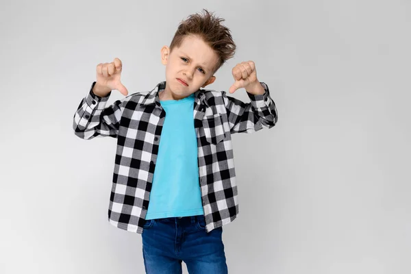 Un beau garçon en chemise à carreaux, chemise bleue et jean se tient sur un fond gris. Le garçon a plié les bras sur sa poitrine. Garçon montrant pouce vers le bas — Photo