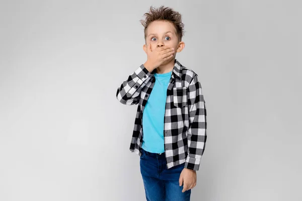 En stilig pojke i en rutig skjorta, ljusblå skjorta och jeans står på en grå bakgrund. Pojken viks armarna över bröstet. Pojken putade ögonen och stänga munnen — Stockfoto