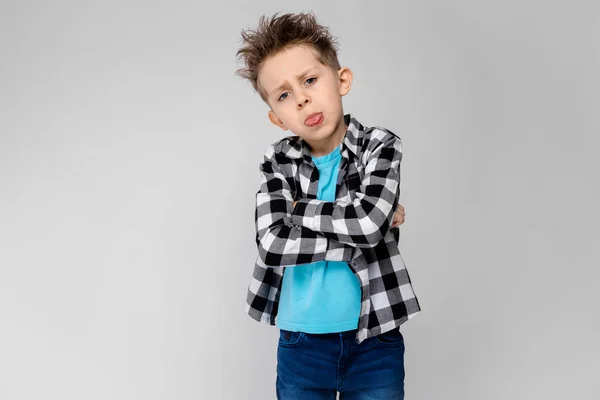 En stilig pojke i en rutig skjorta, ljusblå skjorta och jeans står på en grå bakgrund. Pojken viks armarna över bröstet. Pojken visar språket — Stockfoto