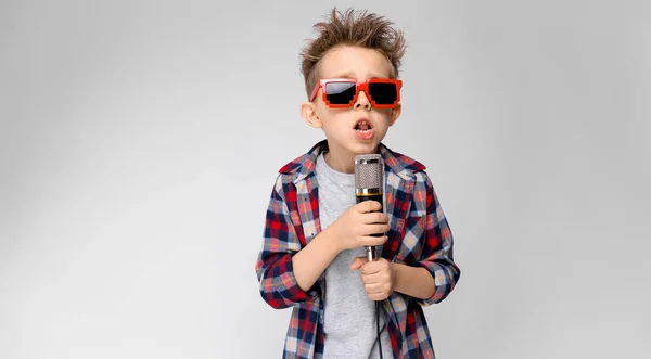 En stilig pojke i en rutig skjorta, grå skjorta och jeans står på en grå bakgrund. En pojke som bär solglasögon. Rödhåriga pojke sjunger i mikrofonen — Stockfoto