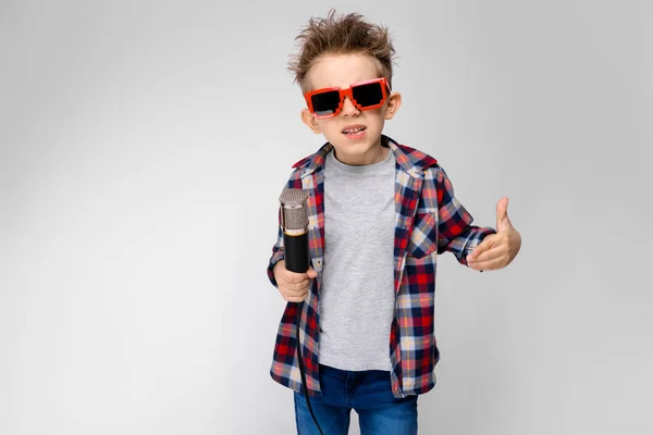 Een knappe jongen in een plaid shirt, grijs shirt en spijkerbroek staat op een grijze achtergrond. Een jongen dragen van een zonnebril. De jongen houdt een microfoon in zijn hand. De jongen is het lezen van rap — Stockfoto