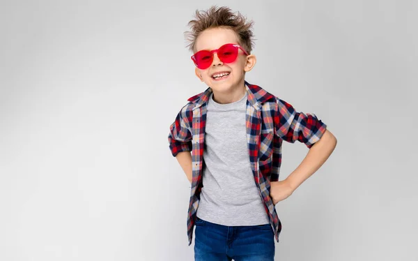 Un chico guapo con una camisa a cuadros, camisa gris y jeans está parado sobre un fondo gris. Un chico con gafas de sol rojas. El chico se puso los puños en las caderas. El chico se ríe. . —  Fotos de Stock