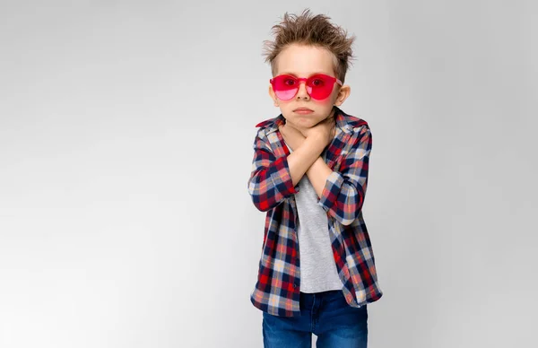 Un chico guapo con una camisa a cuadros, camisa gris y jeans está parado sobre un fondo gris. Un chico con gafas de sol rojas. El niño sostiene sus manos en su garganta . —  Fotos de Stock