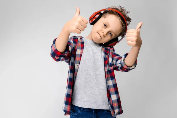 Ένα όμορφο αγόρι σε ένα καρό πουκάμισο, γκρι πουκάμισο και τζιν στέκεται σε γκρι φόντο. Ένα αγόρι στο κόκκινο ακουστικά. Το αγόρι δείχνει το σύμβολο της κατηγορίας. — Φωτογραφία Αρχείου