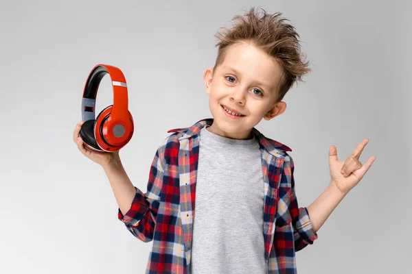 Ένα όμορφο αγόρι σε ένα καρό πουκάμισο, γκρι πουκάμισο και τζιν στέκεται σε γκρι φόντο. Ένα αγόρι στο κόκκινο ακουστικά. Το αγόρι έβγαλε τα ακουστικά του. — Φωτογραφία Αρχείου