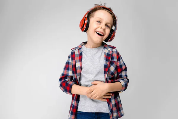 Przystojny chłopak w plaid shirt, szara koszula i jeansy stoi na szarym tle. Chłopiec w czerwonej słuchawki. Chłopiec trzyma ręce na brzuchu. Chłopak śmieje się. — Zdjęcie stockowe