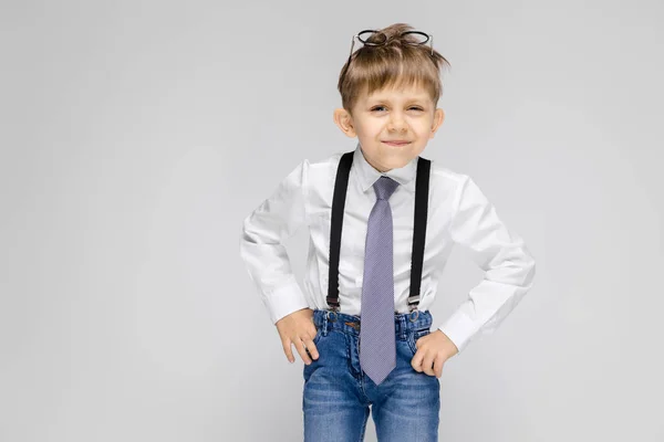 Un niño encantador con una camisa blanca, tirantes, corbata y pantalones vaqueros ligeros se levanta sobre un fondo gris. Gafas en la cabeza — Foto de Stock