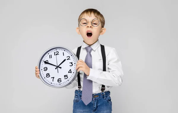 Ein charmanter Junge in weißem Hemd, Hosenträgern, Krawatte und heller Jeans steht vor grauem Hintergrund. der Junge hält eine Armbanduhr — Stockfoto