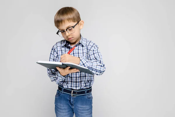 Un niño encantador con una camisa vkletchatoy y pantalones vaqueros ligeros se encuentra sobre un fondo gris. El niño sostiene un cuaderno y un bolígrafo en sus manos . — Foto de Stock
