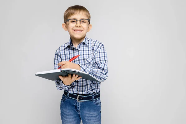 灰色背景下的一个男孩的肖像 一个戴眼镜的男孩 这个男孩手里拿着一本笔记本和一支红色的钢笔 一个男孩在笔记本上做笔记 — 图库照片