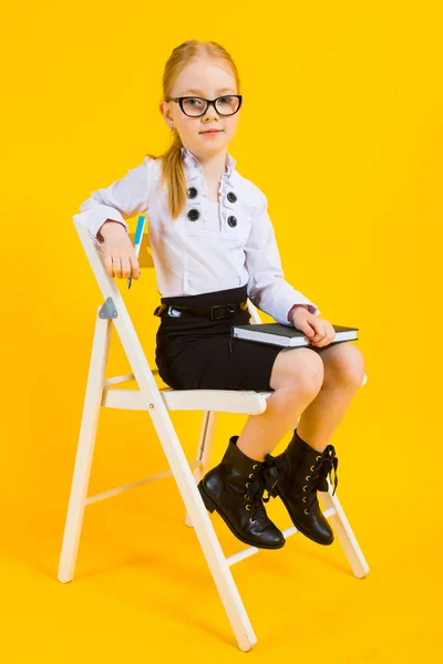 Girl with red hair on a yellow background. A charming girl in transparent glasses sitting on white chair and makes notes in notebook.