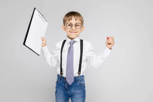 A charming boy in a white shirt, suspenders, a tie and light jeans stands on a gray background. The boy holds a pen and sheets for notes Royalty Free Stock Images