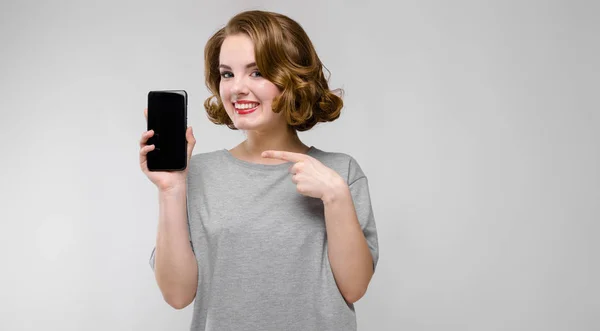 Charmantes junges Mädchen in einem grauen T-Shirt vor grauem Hintergrund. ein Mädchen hält ein Handy in der Hand — Stockfoto