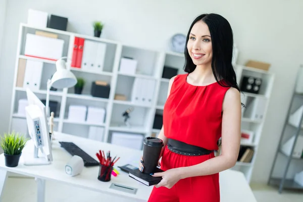 Mooi jong meisje in een rode pak is permanent in het kantoor en houdt een notebook en een glas koffie. — Stockfoto