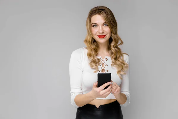 Atractiva joven mujer de negocios rubia en ropa blanca y negra sonriendo sosteniendo el teléfono móvil en sus manos de pie en la oficina sobre fondo gris —  Fotos de Stock