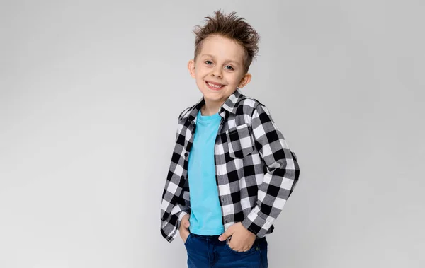 Een knappe jongen in een plaid shirt, blauw shirt en spijkerbroek staat op een grijze achtergrond. De jongen glimlacht en zet zijn handen in de zakken van zijn spijkerbroek — Stockfoto