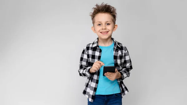 Ein gutaussehender Junge in kariertem Hemd, blauem Hemd und Jeans steht vor grauem Hintergrund. Der Junge hält ein Telefon in der Hand — Stockfoto