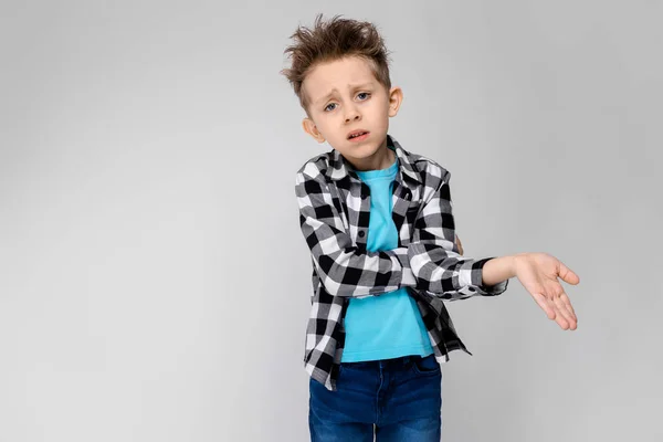 Un chico guapo con camisa a cuadros, camisa azul y pantalones vaqueros está parado sobre un fondo gris. El niño dobló los brazos sobre su pecho. El chico es infeliz. El chico muestra su mano —  Fotos de Stock