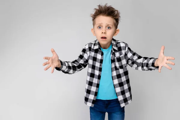 Un chico guapo con camisa a cuadros, camisa azul y pantalones vaqueros está parado sobre un fondo gris. El chico extendió sus manos en ambas direcciones. —  Fotos de Stock