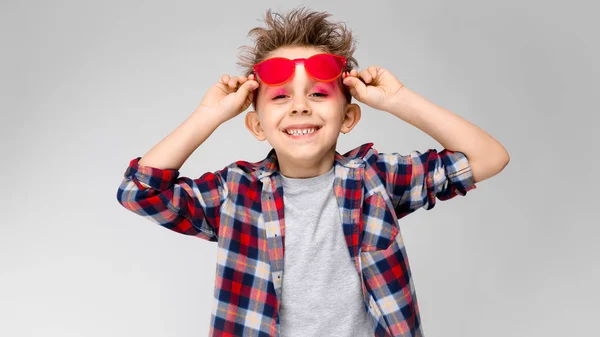 Ένα όμορφο αγόρι σε ένα καρό πουκάμισο, γκρι πουκάμισο και τζιν στέκεται σε γκρι φόντο. Ένα αγόρι σε κόκκινα γυαλιά ηλίου. Το αγόρι κατέχει τα γυαλιά του χέρι — Φωτογραφία Αρχείου