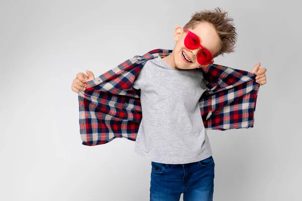 Een knappe jongen in een plaid shirt, grijs shirt en spijkerbroek staat op een grijze achtergrond. Een jongen in rode zonnebril. De jongen trekt zijn shirt terug. — Stockfoto